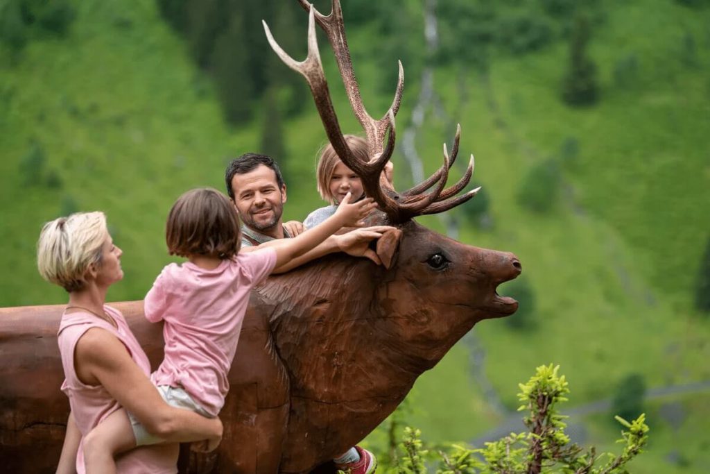 Familie spielt und lacht mit einer Holzskulptur eines Hirsches in der Berglandschaft.