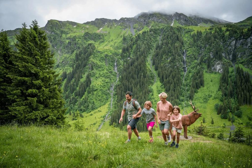 Familie wandert durch eine grüne Berglandschaft mit einem Hirsch im Hintergrund.
