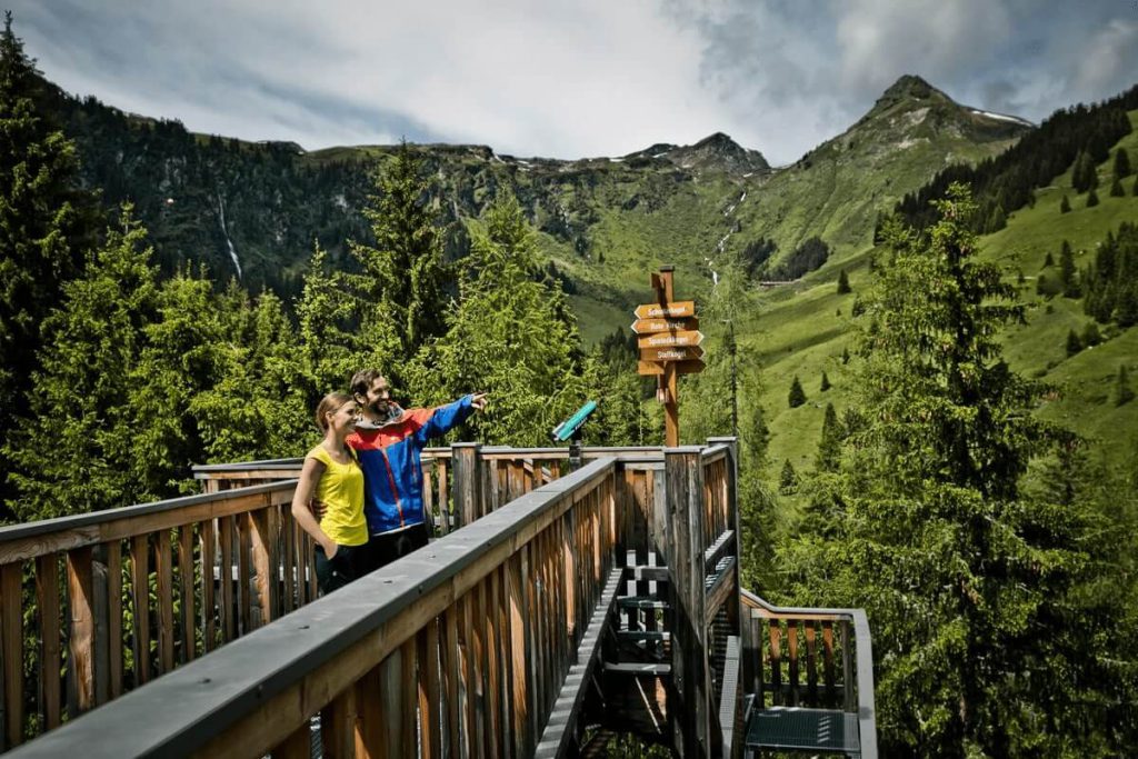 Paar auf Aussichtsplattform in den Bergen mit Wegweiser und grüner Landschaft.