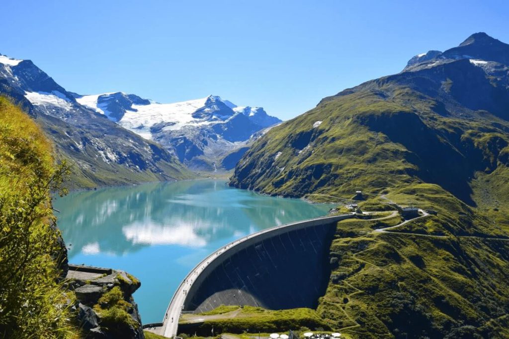 Blick auf einen klaren Stausee mit schneebedeckten Bergen und grünen Wiesen im Sommer.