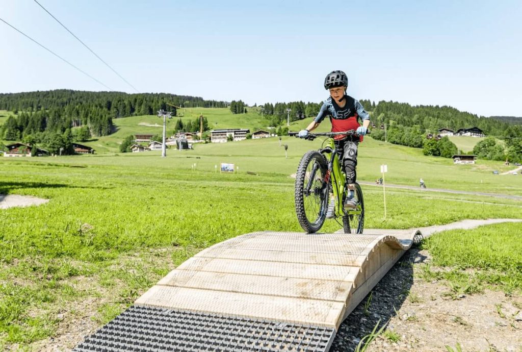 Junge fährt mit dem Mountainbike über eine Holzrampe in der grünen Berglandschaft.