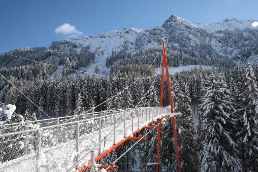 Schneebedeckte Berglandschaft mit einer Hängebrücke zwischen hohen Tannen im Winter.