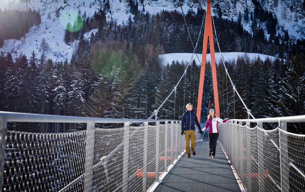 Paar spazier auf einer Hängebrücke in verschneiten Bergen, umgeben von winterlicher Natur.