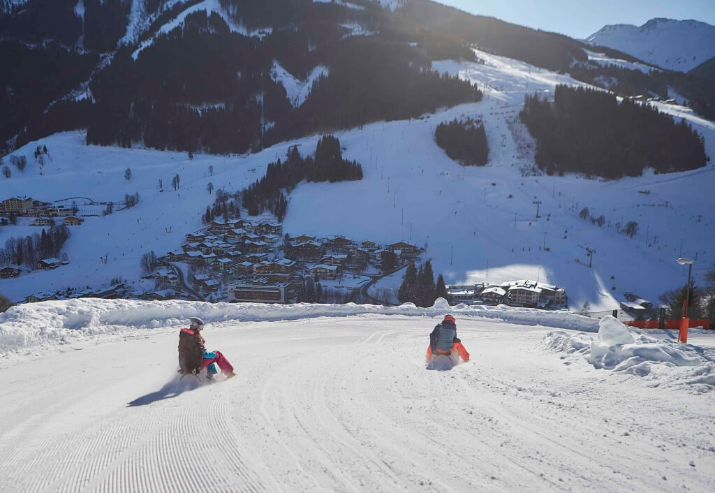 Kinder rodeln auf einer verschneiten Piste in Saalbach-Hinterglemm mit Bergpanorama.